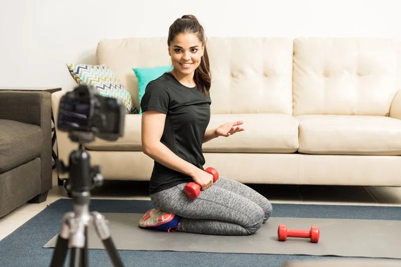 A woman vlogging for a fitness channel, kneeling on a yoga mat with small hand weights.