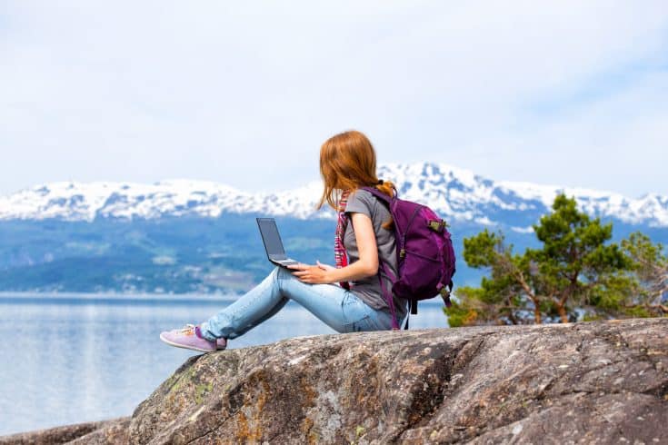 A woman sits on a rock and blogs on a laptop computer; in the background is a snow-capped mountain range.