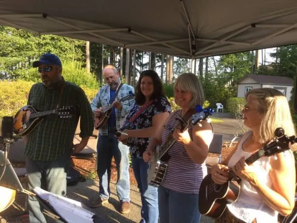 A group plays instruments outdoors beneath a pop up gazebo, with woods in the background.