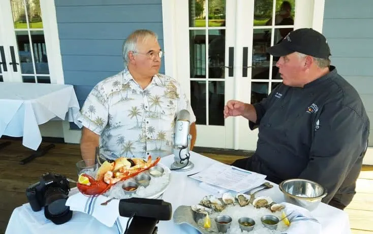 Chef Dennis recording an interview. On the table between them are various seafood dishes.