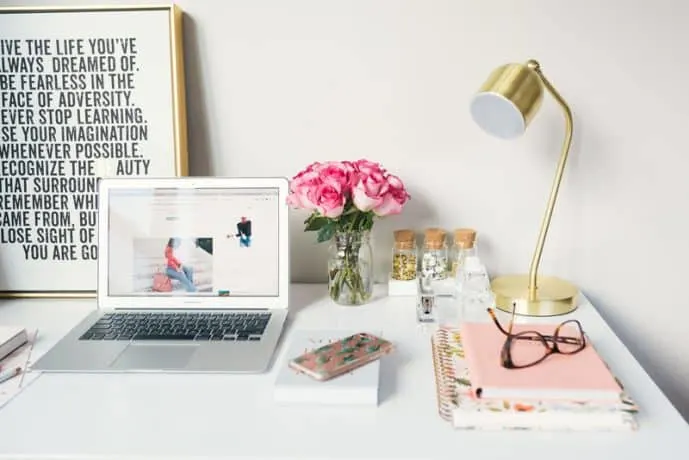 A desktop with a laptop computer, smart phone, bouquet of flowers, lamp, notebooks and a pair of glasses on the surface.