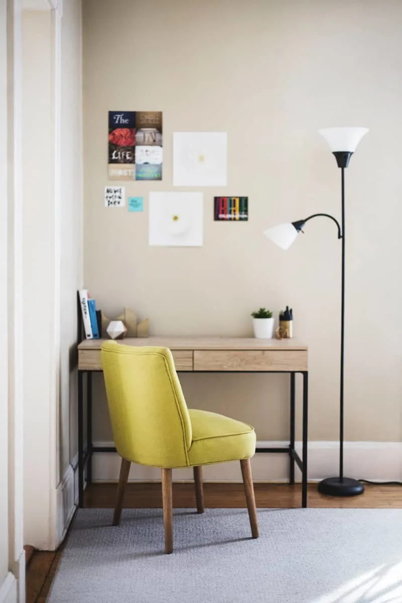 A desk sits in the corner of a light with plenty of natural light. A green upholstered chair sits in front of it.