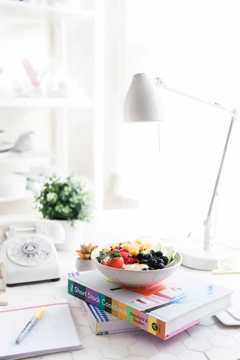 A desk with a lamp, stack of books topped with a bowl of fruit, phone, plant, and planner with pen.