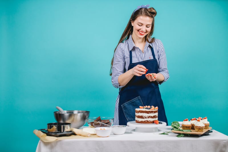 A woman standing in front of a teal background, working on decorating a carrot cake.