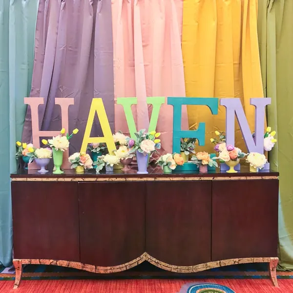 A Haven photo booth, showing a rainbow of colored curtains in the background, and a table top decorated with colorful vases and multi-colored wooden letters reading "Haven".