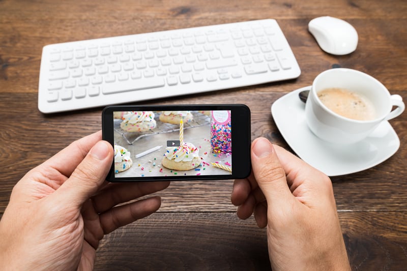A smart phone plays a video ad; in the background is a tabletop with wireless keyboard and mouse, and a cup of coffee.