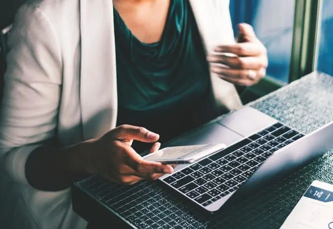 A woman scrolls through her smartphone while working at a laptop computer.