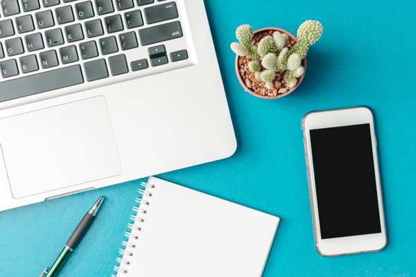 A laptop computer keyboard, a pen, a notebook, a smart phone, and a small potted cactus all sit on a teal background.
