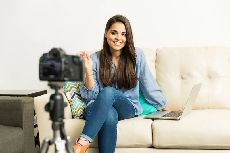 A woman sits on a white couch with a laptop beside her, vlogging.