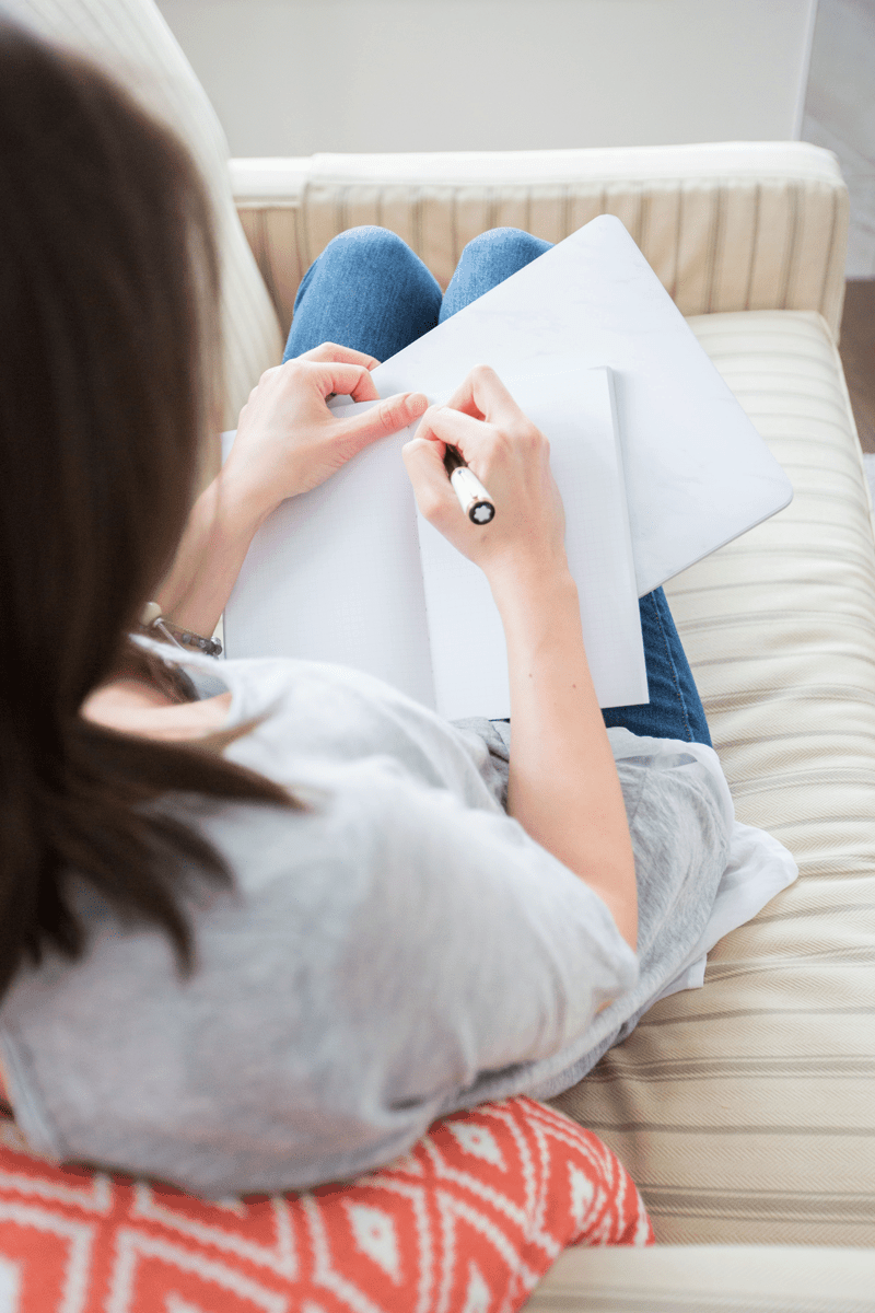 A woman writes in a notebook while laying back on a couch.