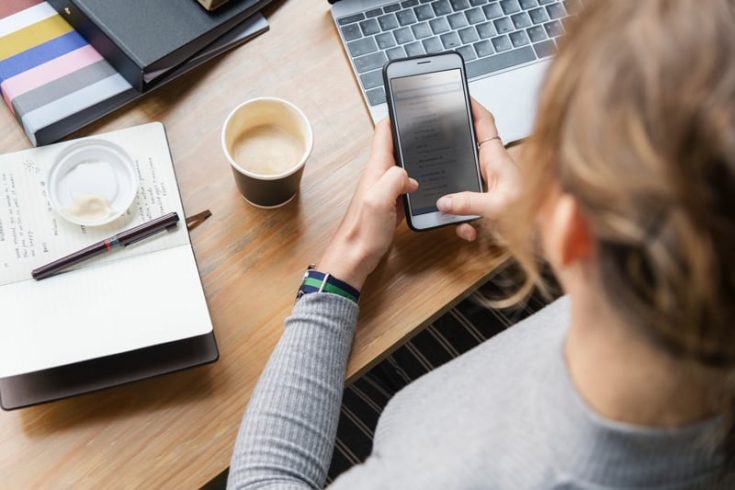 A woman holding a smart phone with a laptop, coffee, and notebook in the background on a table top.