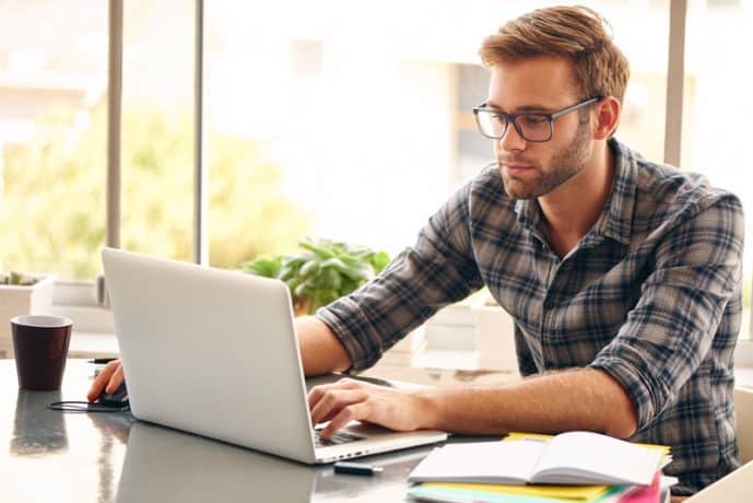 A man works on a laptop computer, with an open notebook and cup of coffee beside it.