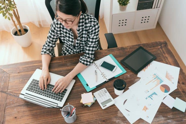 A woman typing on a laptop. On the same tabletop are a notebook, a tablet, and several charts and graphs.