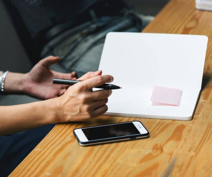 hands holding a pen over a blank notebook next to a smartphone 