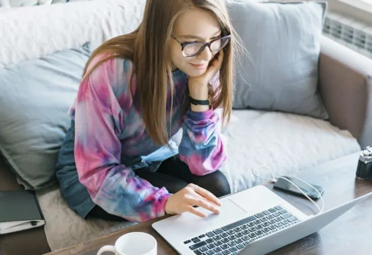 A woman wearing a tie dye shirt sits on a couch and uses a computer.