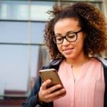 woman with glasses, outside, looking down at her phone and smiling
