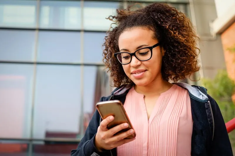 woman with glasses, outside, looking down at her phone and smiling