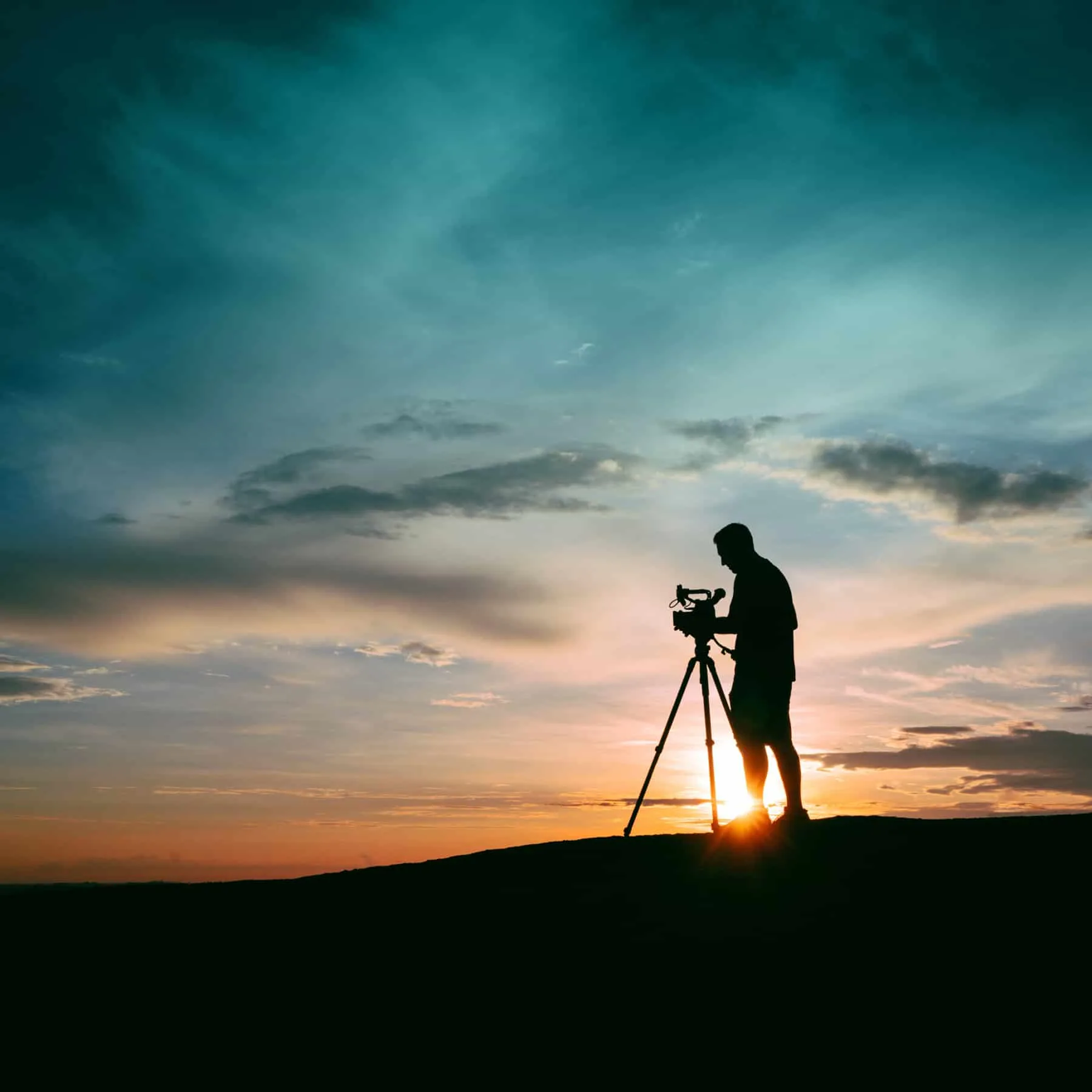 A man in silhouette setting up a camera, with the sun setting behind him.