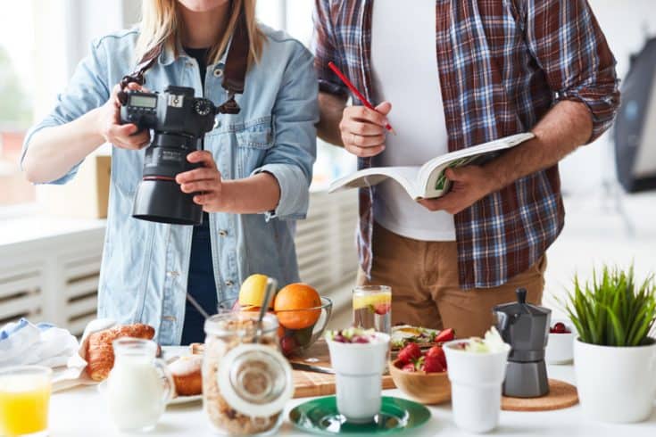 A woman taking photographs of food, while a man writes notes in a notebook beside her.