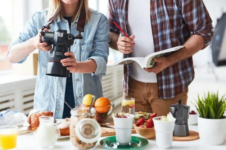 A woman takes photos of food, while a man takes notes in a notebook beside her.