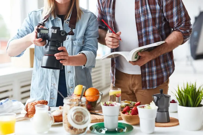 woman with camera pointing down at a table of food, man standing next to her with an open notebook