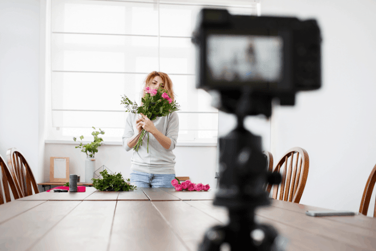 A camera records a woman holding a bouquet of flowers across a kitchen table.