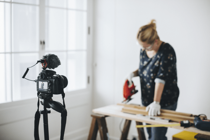 A woman films a DIY video, cutting wood with a jigsaw.