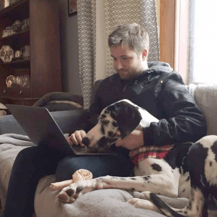 Alex Roth works on a laptop computer. A spotted Great Dane lays at his side, watching him work.