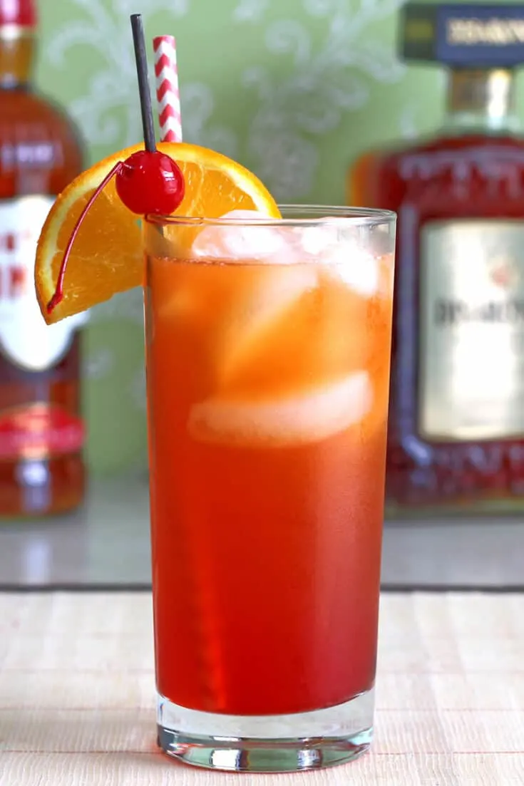 A red-orange cocktail, garnished with striped straw, orange slice and cherry.