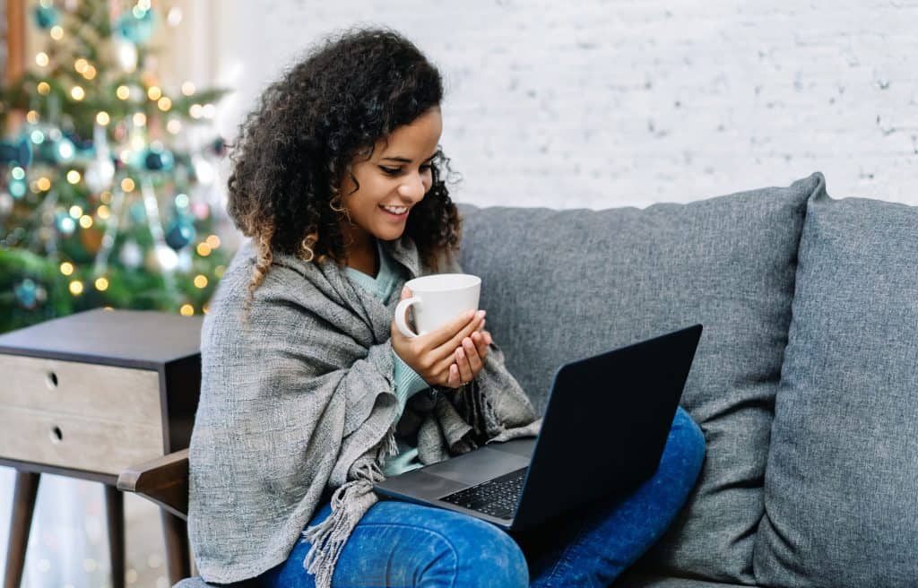 Woman on couch with scarf wrap, holding a mug, looking at laptop