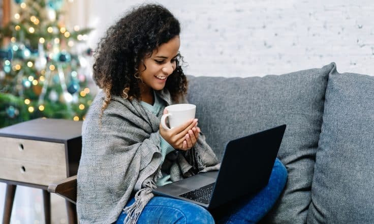 Woman on couch with scarf wrap, holding a mug, looking at laptop
