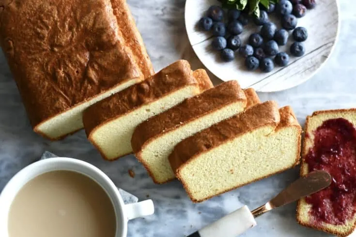 A loaf of bread, sliced. A cup of coffee and plate of blueberries sits to the side. One slice of bread is being spread with jam.