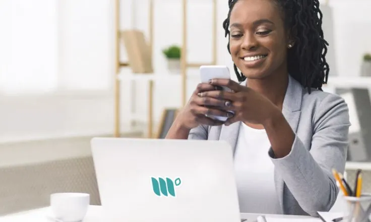 woman using phone sitting next to laptop at a desk