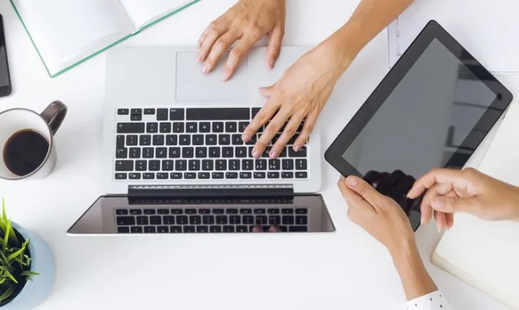 above view of 1 pair of hands typing on a laptop and another holding an ipad