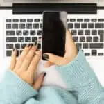 the hands of a woman in a light blue sweater using a smartphone while holding a laptop in their lap