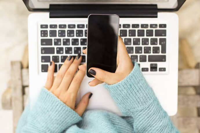 the hands of a woman in a light blue sweater using a smartphone while holding a laptop in their lap