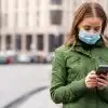 woman in a green jacket wearing a mask and looking at her phone on a city street