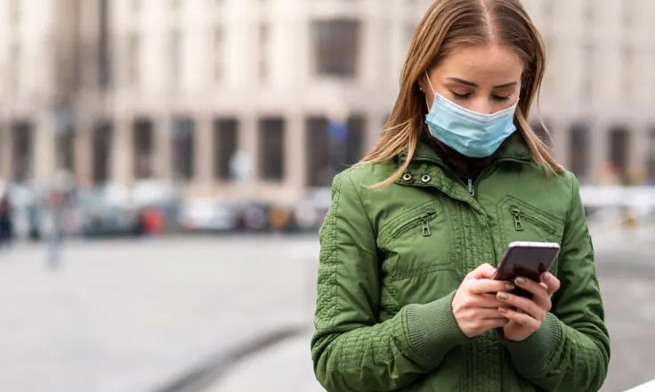 woman in a green jacket wearing a mask and looking at her phone on a city street