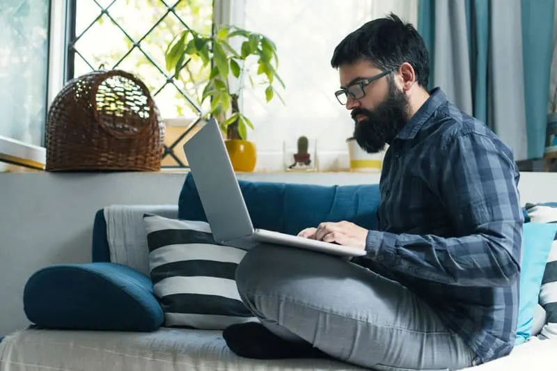 man typing on a laptop in his lap while sitting on a gray couch near a window