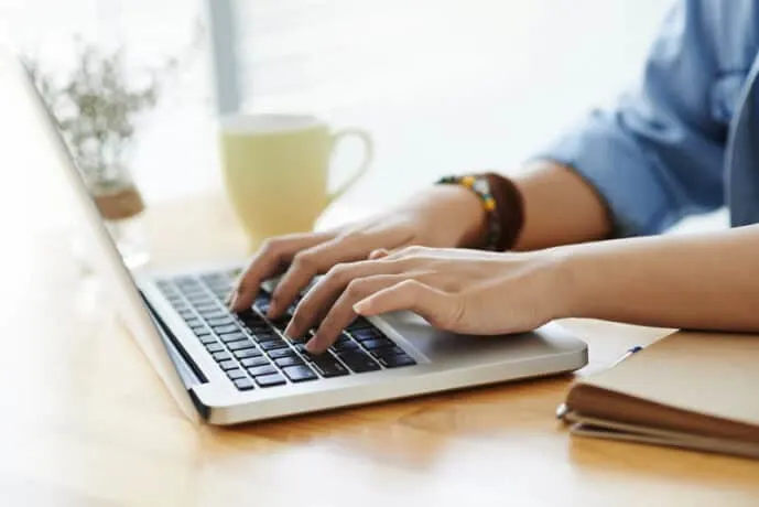 person drinking coffee and typing on a laptop on a wooden desk