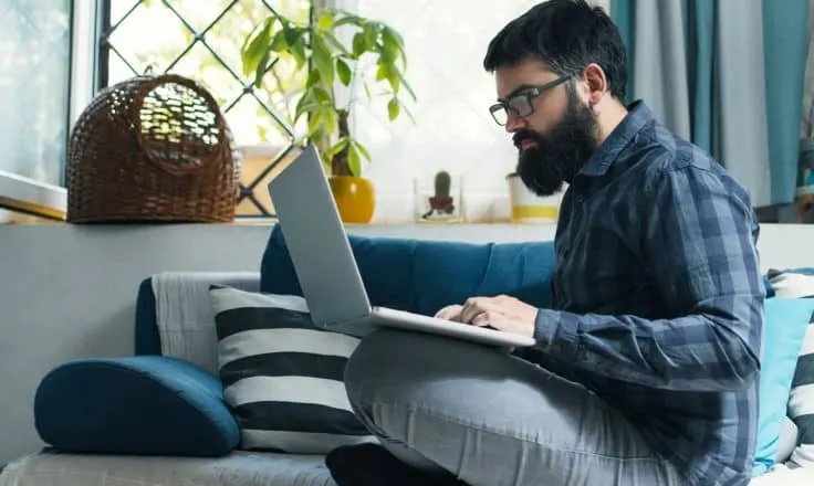male developer wearing plaid shirt coding on a laptop from a couch