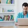 male blogger in blue shirt writing on a laptop from his home office with bookshelf behind him
