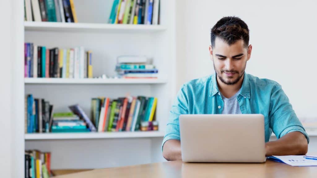 male blogger in blue shirt writing on a laptop from his home office with bookshelf behind him