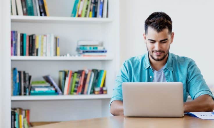 male blogger in blue shirt writing on a laptop from his home office with bookshelf behind him