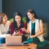 three female bloggers looking at a smartphone and a laptop together on a couch