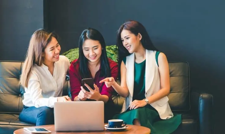 three female bloggers looking at a smartphone and a laptop together on a couch