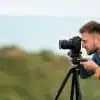 male travel blogger taking photos on a DSLR camera on a tripod with cliffs and ocean behind him