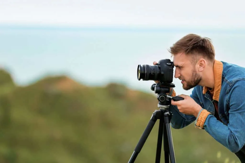 male travel blogger taking photos on a DSLR camera on a tripod with cliffs and ocean behind him