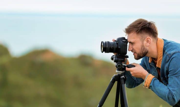 male travel blogger taking photos on a DSLR camera on a tripod with cliffs and ocean behind him