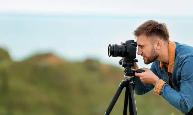 male travel blogger taking photos on a DSLR camera on a tripod with cliffs and ocean behind him